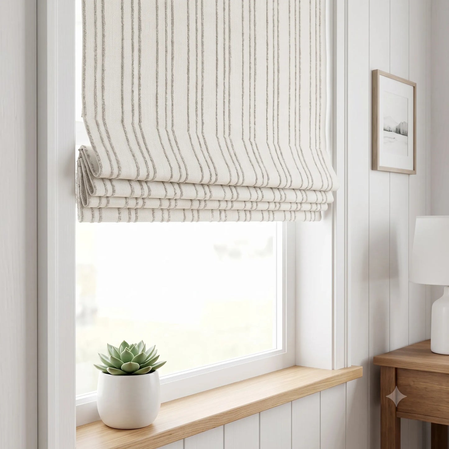 A Roman blind with vertical stripes in cream and gray, partially drawn over a window in a bright, modern room with a wooden sill, a small potted succulent, and a wooden side table with a lamp.