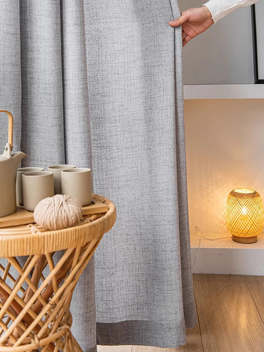 Person holding a gray curtain next to a wooden side table with ceramic items and a lamp.