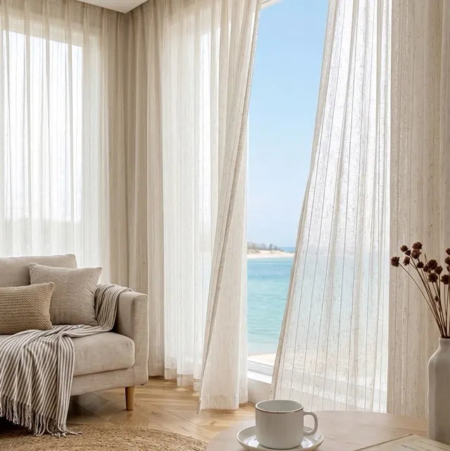 Living room with beige sofa, white curtains, and a view of the ocean.