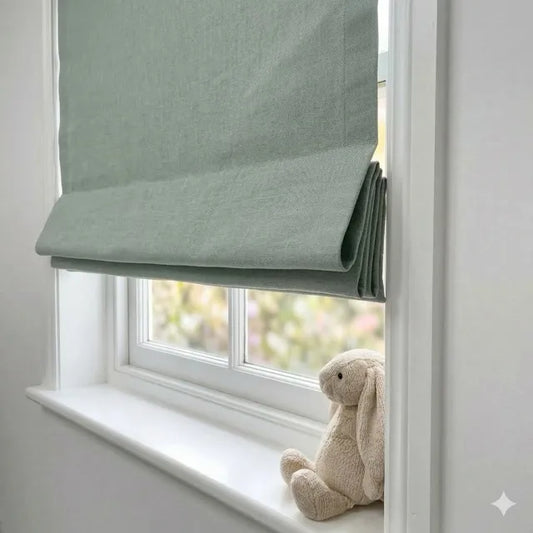 Light green roman blind partially open on a window with a teddy bear on the windowsill.
