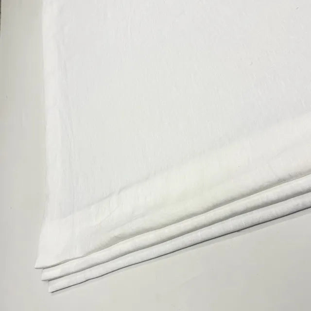 A close-up of folded white linen fabric, likely cotton or linen, showing its soft texture and layered edges against a light neutral background.
