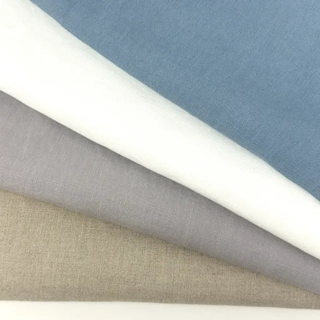 four folded french linen fabric fabric swatches in natural, blue, white, and light gray, layered diagonally on a white surface.