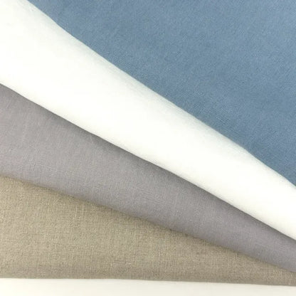 four folded french linen fabric fabric swatches in natural, blue, white, and light gray, layered diagonally on a white surface.