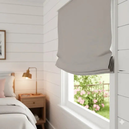 A light gray fabric Roman blind partially drawn over a window in a bright, modern bedroom with white shiplap walls and a wooden nightstand.