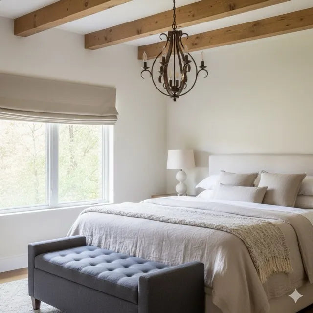 A styled bedroom interior featuring a neatly made bed with neutral linens, a dark tufted bench at the foot, a black metal chandelier, exposed wooden ceiling beams, and a large window with a washed french linen natural color roman shade.