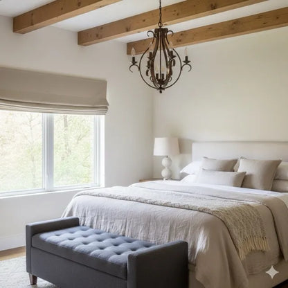 A styled bedroom interior featuring a neatly made bed with neutral linens, a dark tufted bench at the foot, a black metal chandelier, exposed wooden ceiling beams, and a large window with a washed french linen natural color roman shade.