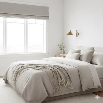 A minimalist bedroom featuring a bed dressed in light beige and off-white bedding with a textured fringed throw blanket. The room has white walls, a large window with a gray french linen roman shade, a brass wall sconce, and a small side table with a vase of flowers.