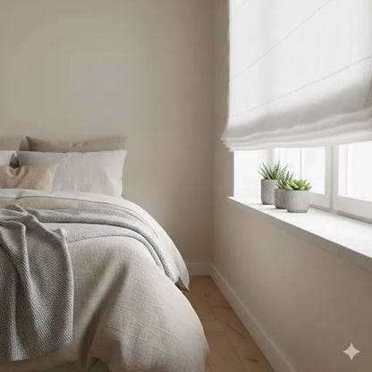 A softly lit bedroom corner with a bed dressed in neutral beige and white bedding, a textured gray throw blanket, a white window blind, and two small potted succulents on the windowsill.