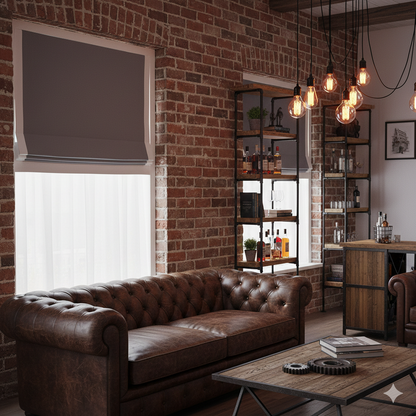 Cozy living room with grey roman shade with white trims, brown leather sofa, brick wall, and wooden shelves.