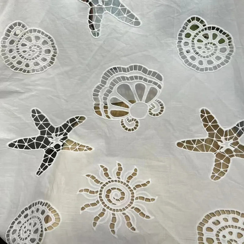White eyelet lace fabric featuring embroidered seashell and starfish motifs in black and metallic gold.