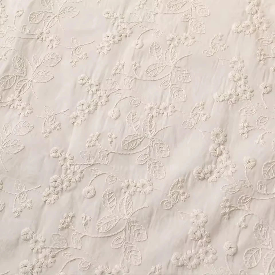 Close-up of a cream-colored fabric with delicate floral embroidery and embroidered leaf patterns.