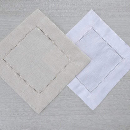 Two square cotton napkins with embroidered borders, one in light beige and one in white, arranged on a light gray wood-textured surface.