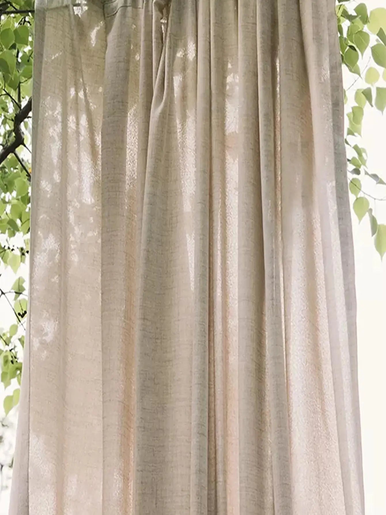 Beige curtain with natural light filtering through, surrounded by green leaves.
