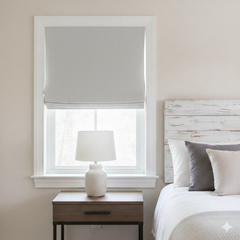 Window with gray roller blind, white lamp on nightstand, and bed with pillows in a bedroom setting.