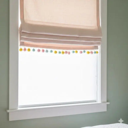Window with pink roman shade and colorful pom-pom garland on a light gray wall.