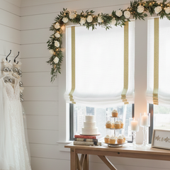 A room with white relaxed roman shade with gold glitter trim with Wedding dress hanging on a rack and wedding cake in the background.