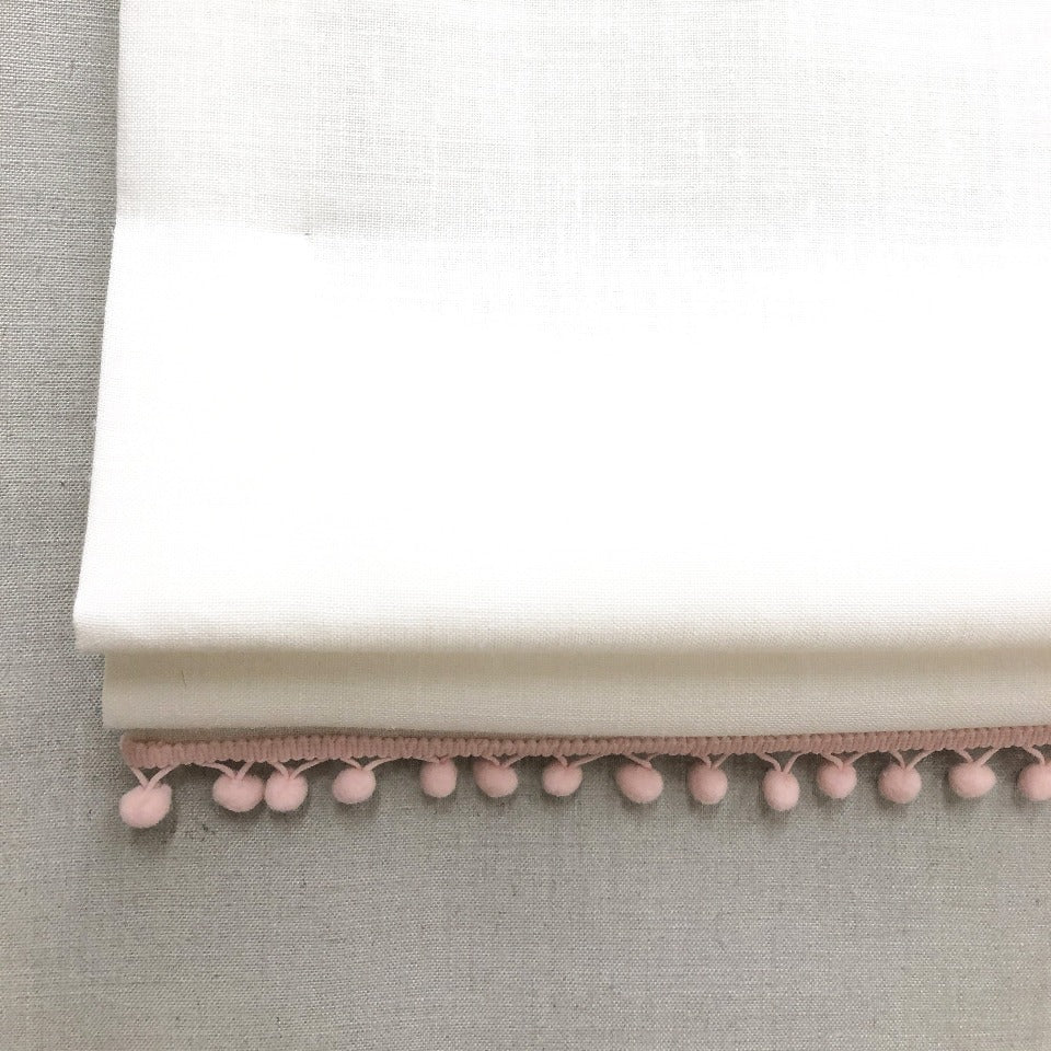 White linen flat relaxed Roman shade featuring soft pink pom pom trim, ideal for nursery or kids' room decor with optional blackout lining.