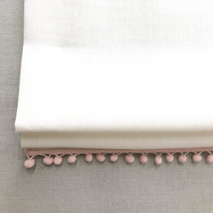 White linen flat relaxed Roman shade featuring soft pink pom pom trim, ideal for nursery or kids' room decor with optional blackout lining.